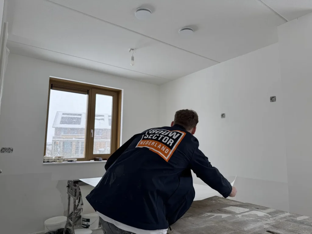 Behanger van Bouwsector Nederland in donkerblauwe jas pakt een baan vlies van de plakmachine in een lege nieuwbouwkamer